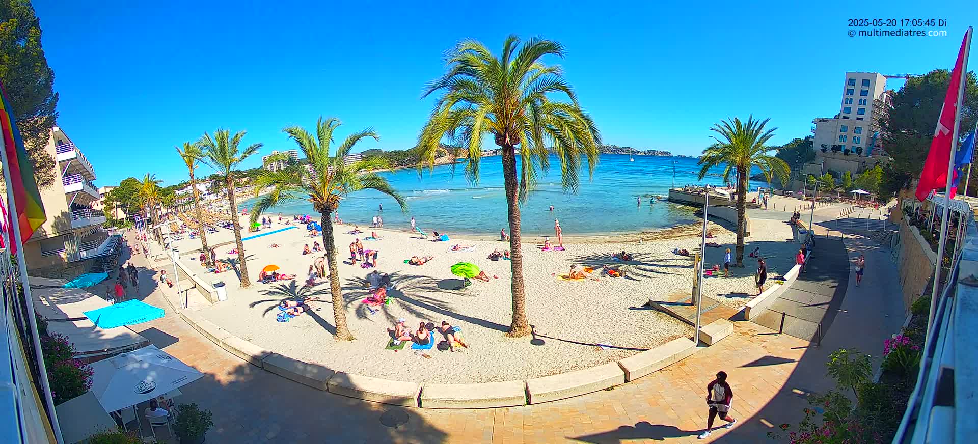 A sunny beach scene shows a sandy shore with people sunbathing and swimming in calm, clear water, palm trees lining the beach and a walkway leading to nearby buildings.