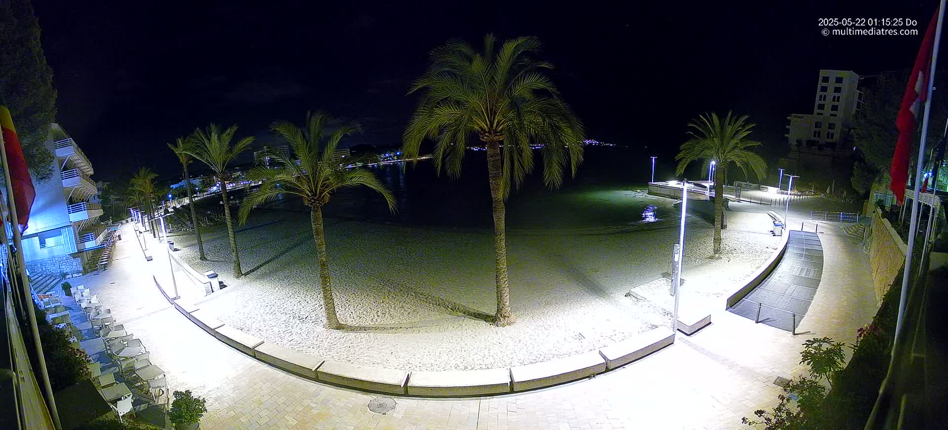 A nighttime, overhead view of a sandy beach, palm trees, and a paved walkway illuminated by lights, with a calm body of water visible in the background.