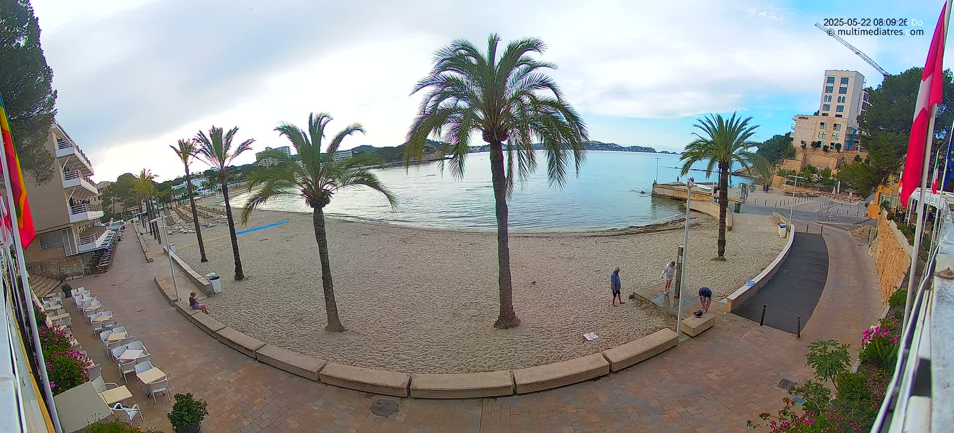 A sandy beach with palm trees and a few people is visible from an elevated vantage point, overlooking a calm body of water under a partly cloudy sky.