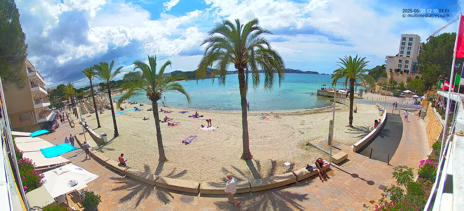 A sandy beach with sunbathers is seen from an elevated viewpoint on a partly sunny day, with palm trees lining the shore and buildings visible in the background.