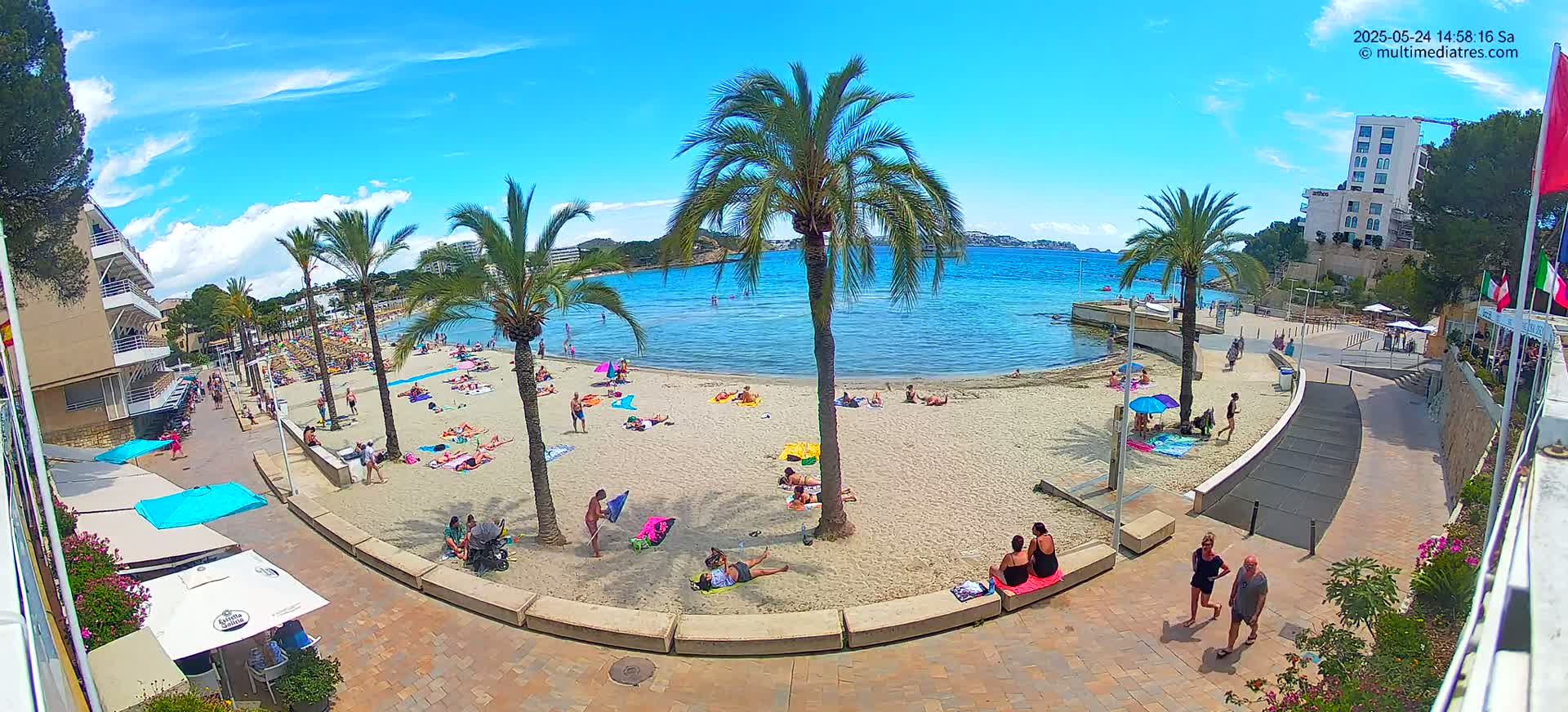 A sunny beach scene shows people relaxing on a sandy beach next to calm, clear blue water under a mostly sunny sky, with palm trees and buildings in the background.