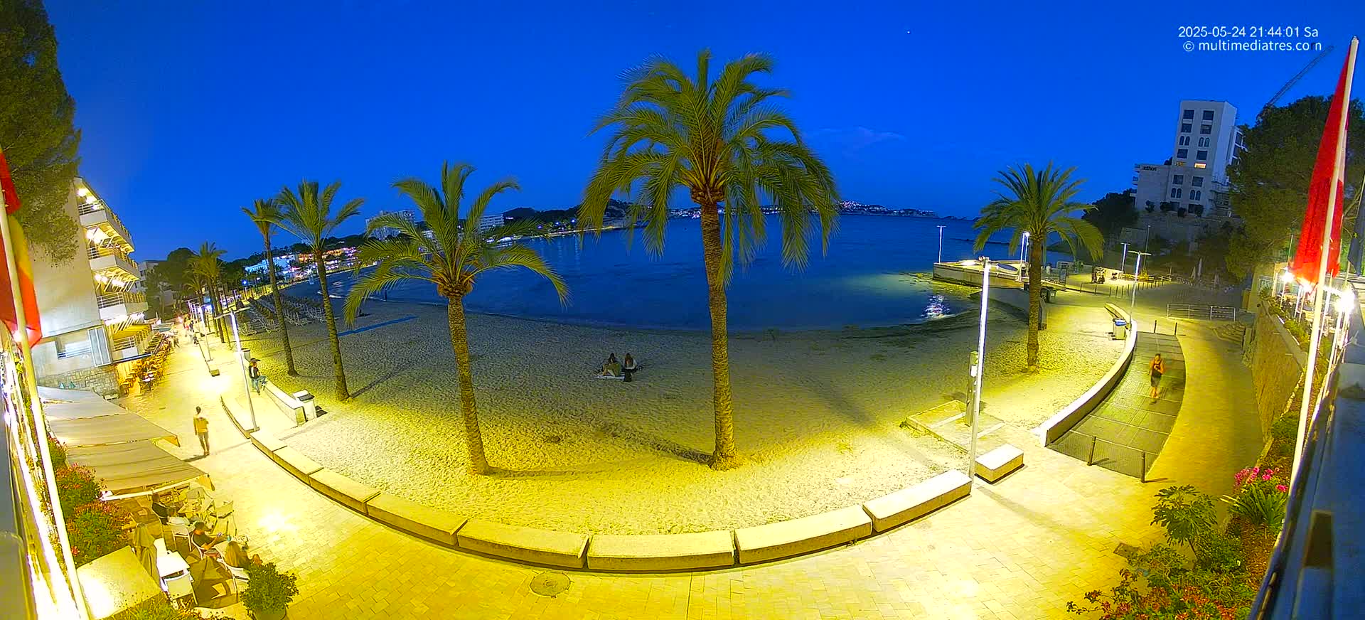 A sandy beach at night, illuminated by artificial lights, is bordered by a paved walkway lined with palm trees, buildings, and outdoor seating areas, under a clear, dark blue sky.
