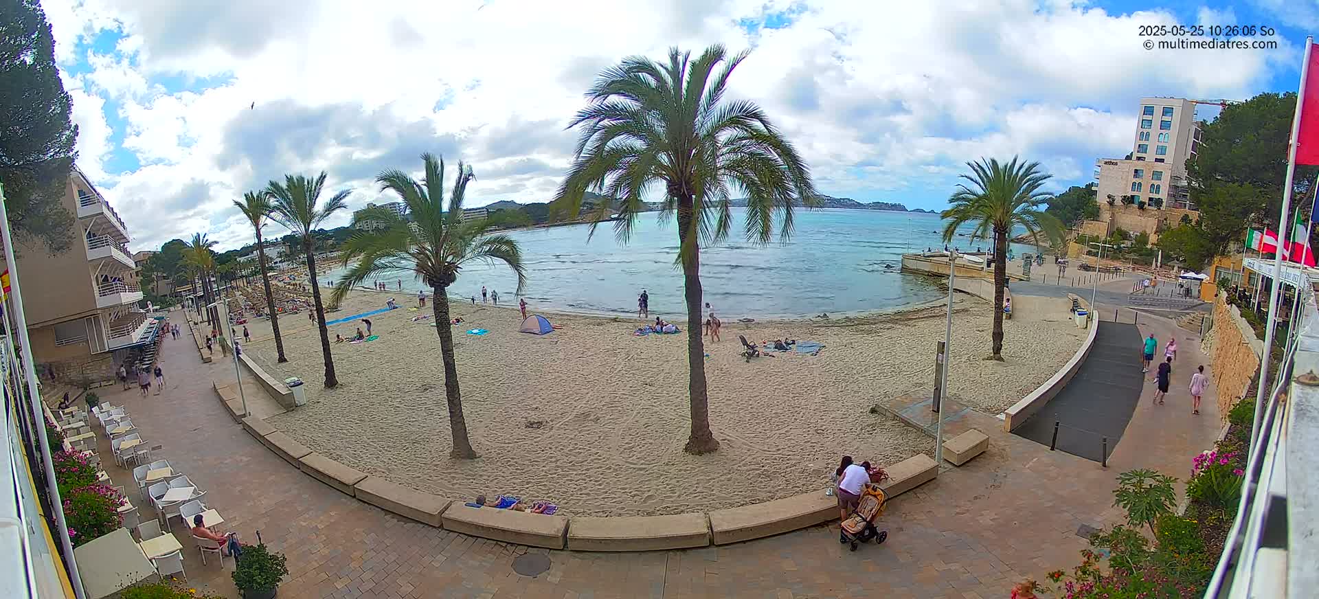A partly cloudy day at a sandy beach with palm trees, people sunbathing and walking along a paved walkway, and a calm sea in the background.