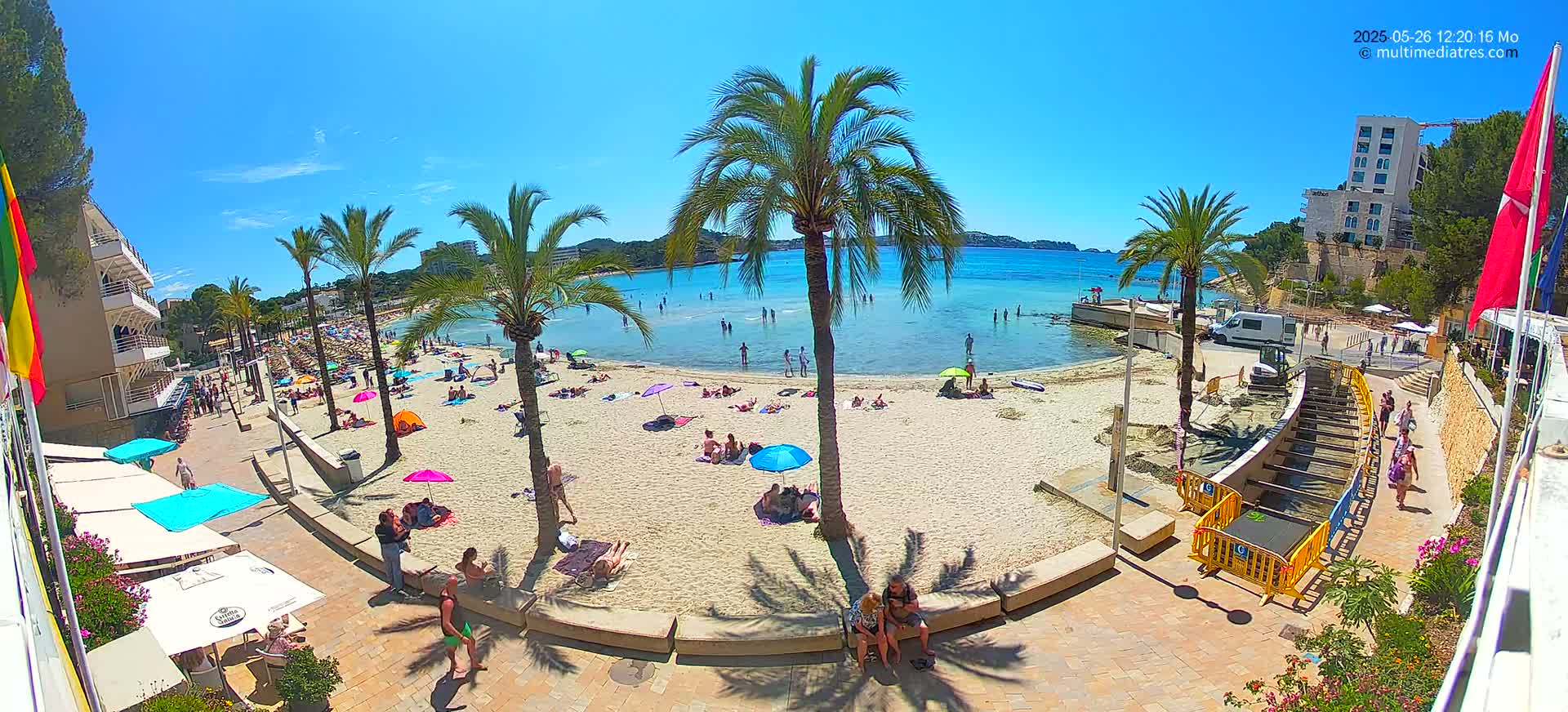A sunny beach scene shows numerous people relaxing on the sand and in the shallow water, under palm trees and umbrellas, near buildings and a walkway.