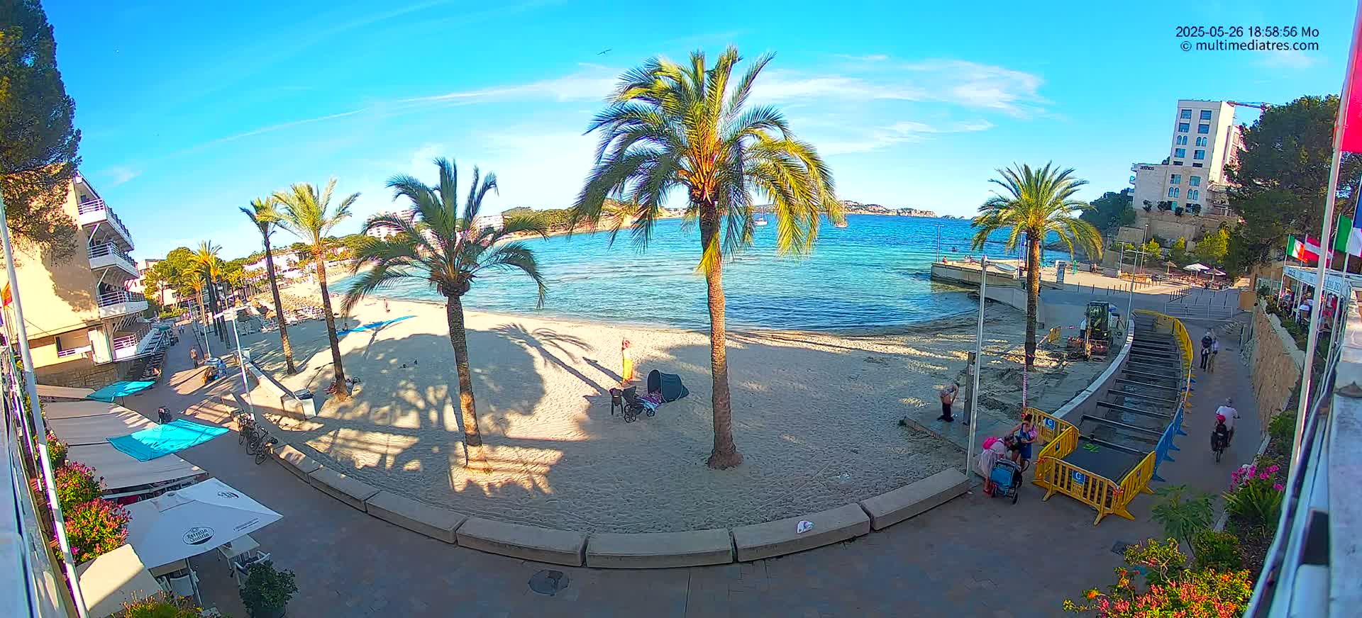 A sunny beach scene shows a sandy beach with palm trees, calm turquoise water, and buildings along the shore, with some people relaxing on the sand and others walking along the paved path.