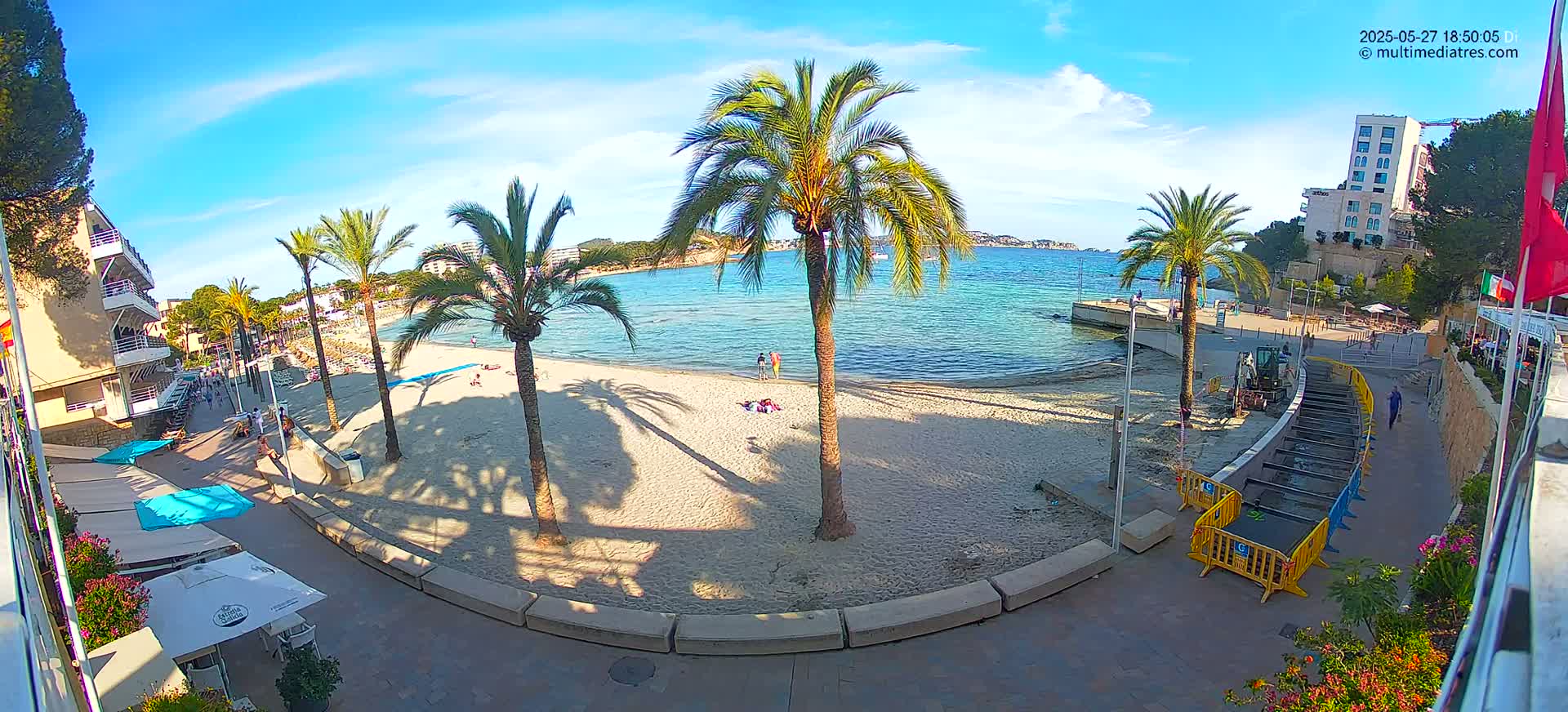 A sandy beach with palm trees and buildings is visible under a sunny sky, with some construction or maintenance equipment present on the paved walkway.