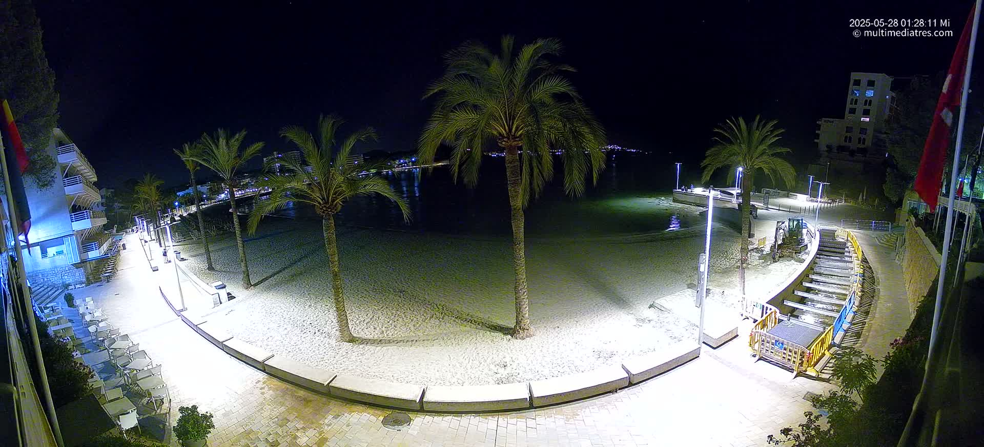 A nighttime aerial view of a sandy beach bordered by palm trees, buildings, and a paved walkway with outdoor seating, showing some construction work near the water.
