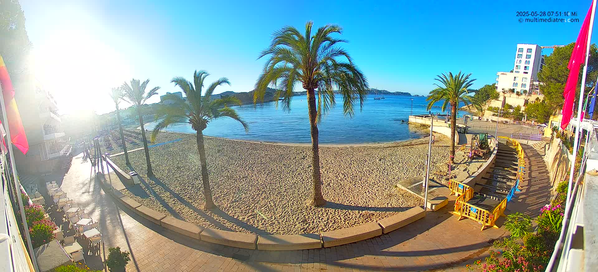 A sandy beach with palm trees and a calm, blue bay is viewed from an elevated position on a sunny day, with construction or maintenance occurring on a walkway leading down to the beach.