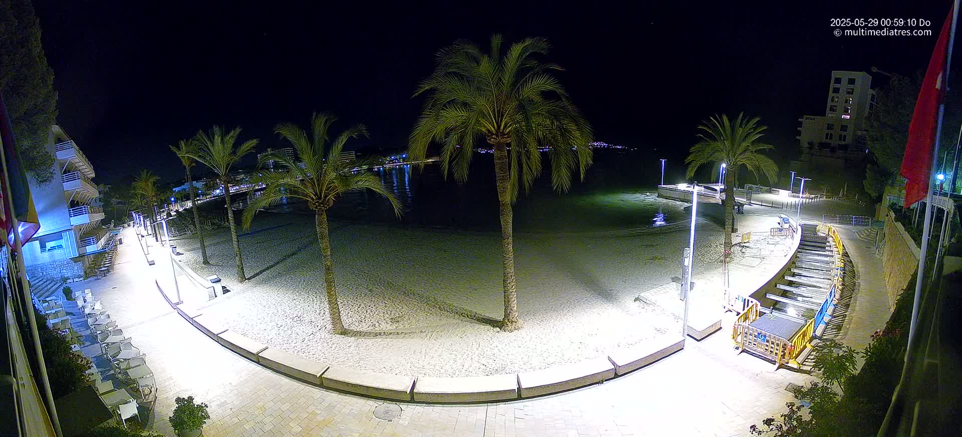 A nighttime aerial view of a sandy beach bordered by a paved walkway, palm trees, and buildings, under a clear, dark sky.