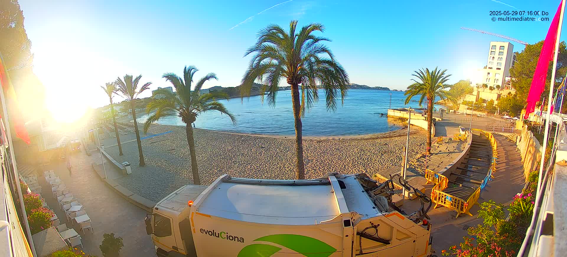 A sunny day at a beach with palm trees, a garbage truck, and a building in the background.