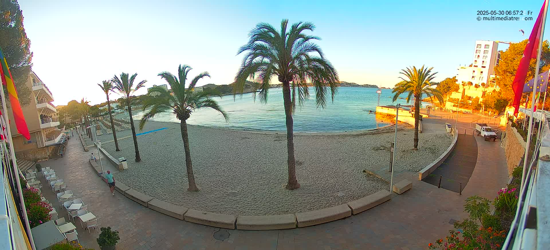 A sandy beach with calm water, palm trees, and buildings is visible under a clear, bright sky.