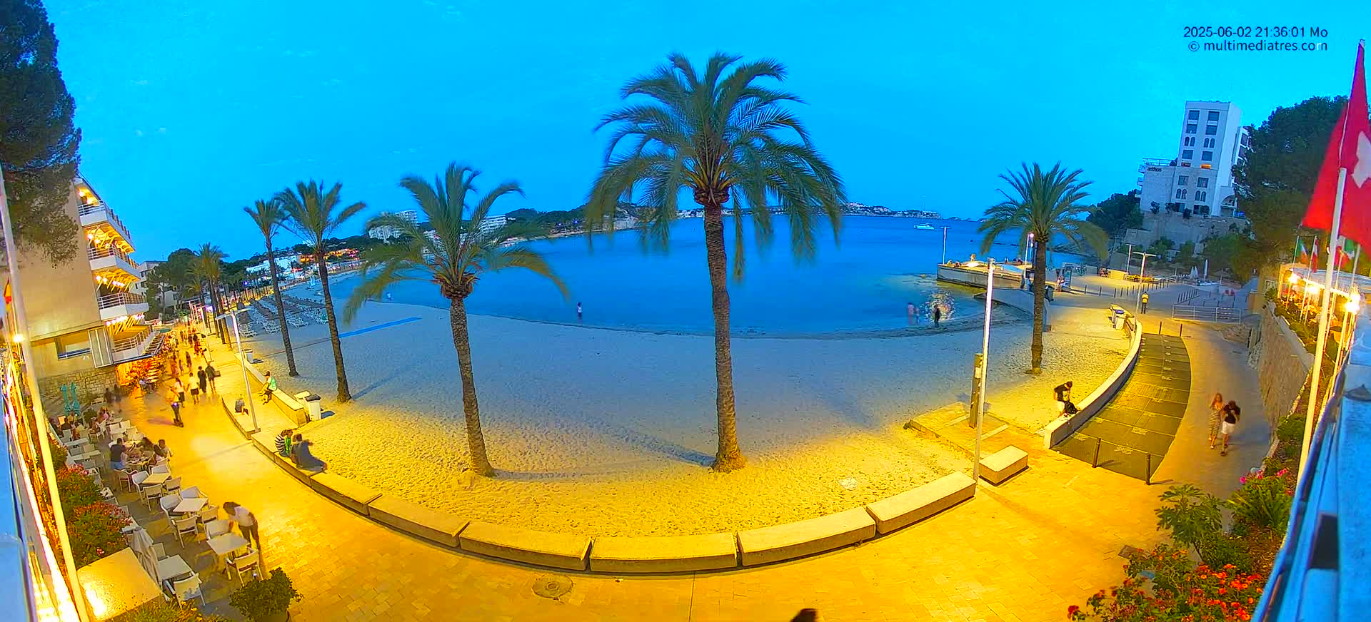 A sandy beach at dusk, illuminated by artificial lights, is bordered by a paved walkway lined with palm trees, buildings, and outdoor seating areas, with a calm body of water visible beyond.