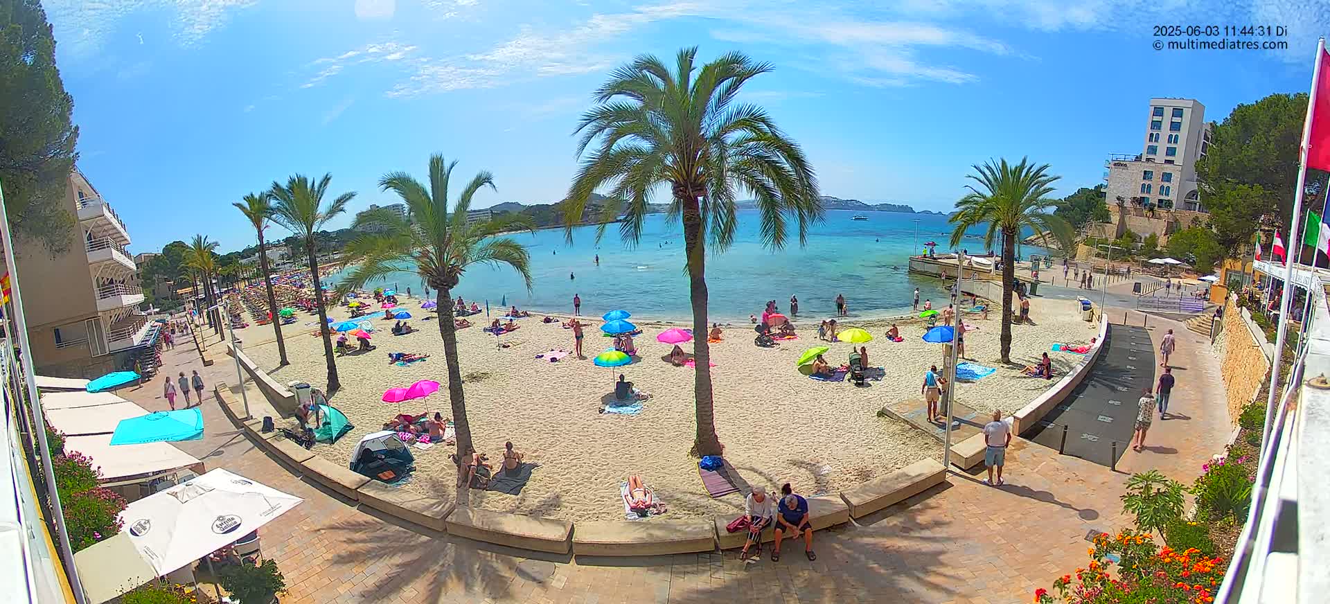 A sunny beach scene with palm trees, colorful umbrellas, and people relaxing on the sand next to a calm, clear body of water.