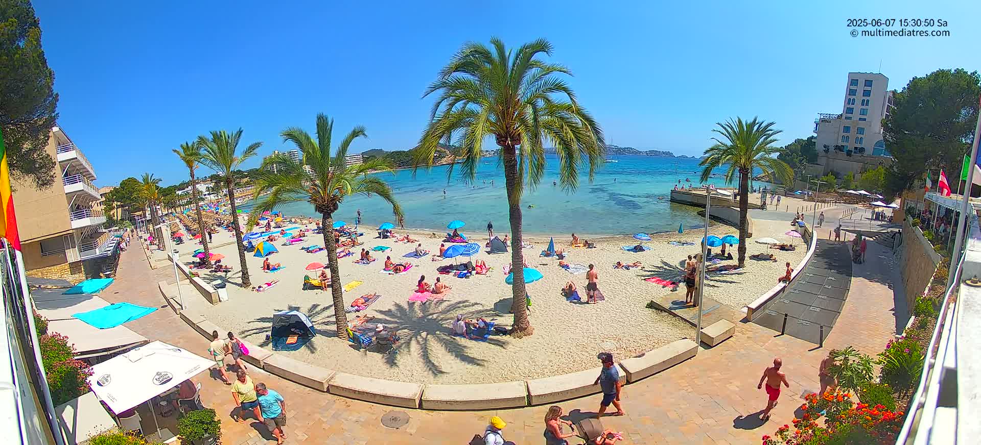A sunny beach scene shows many people relaxing on a sandy beach under colorful umbrellas near palm trees and turquoise water, with buildings and walkways visible.