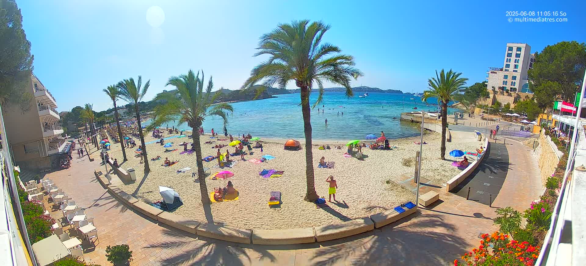 A sunny beach scene shows people relaxing on the sand under colorful umbrellas, with palm trees lining the shore and buildings visible beyond the sandy area.