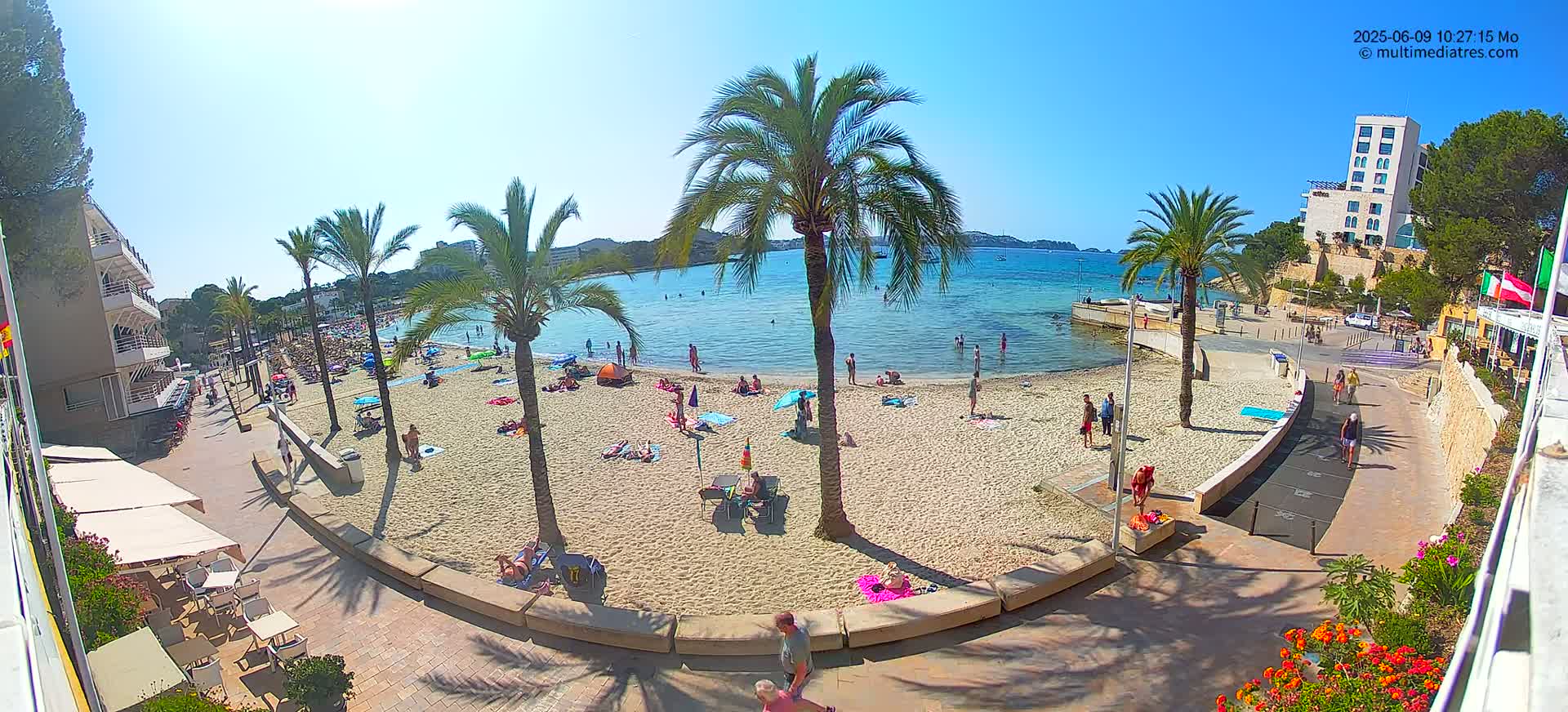 A sandy beach with numerous people sunbathing and swimming, bordered by palm trees, buildings, and a paved walkway, under a clear blue sky.