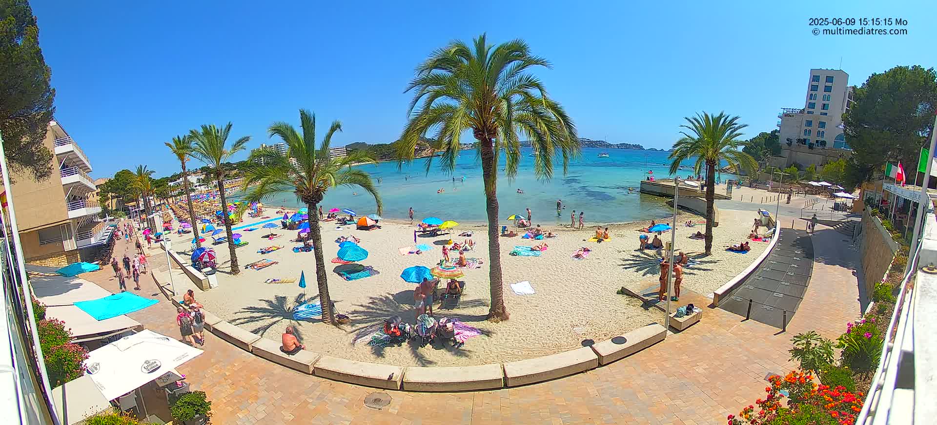 A sunny beach scene shows people relaxing under colorful umbrellas on a sandy beach next to calm, clear water, with palm trees and buildings visible in the background.