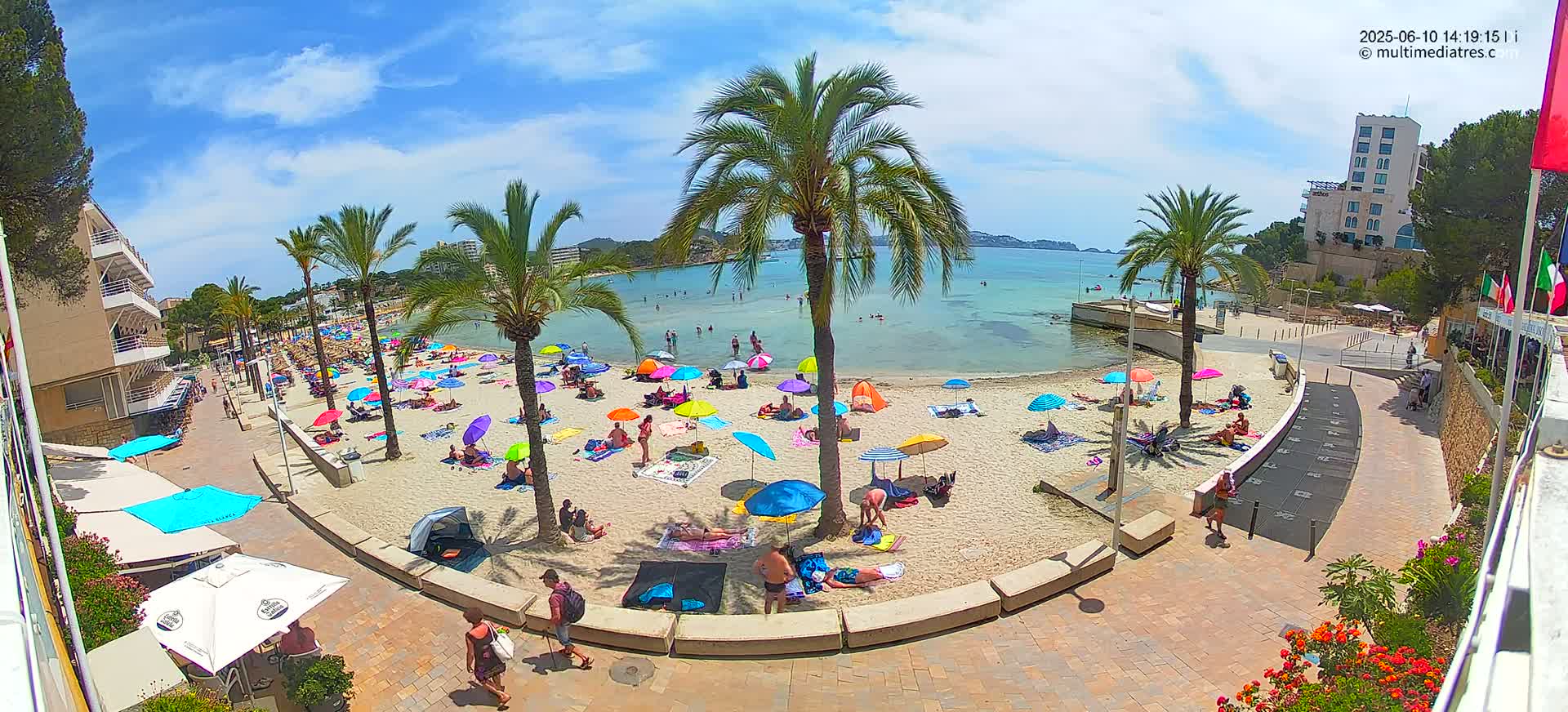 A sandy beach with numerous colorful umbrellas and sunbathers is situated next to a paved walkway lined with palm trees and buildings, all under a sunny sky.