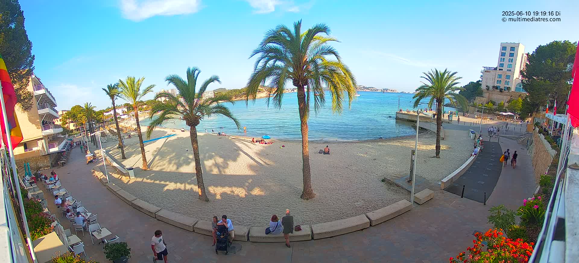 A sandy beach with palm trees and several people, some sitting at outdoor cafes, is situated next to a calm blue bay under a clear, sunny sky.