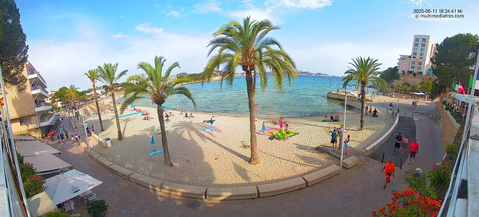 A sandy beach with palm trees and people relaxing under a sunny sky, next to a paved walkway leading to buildings and restaurants.