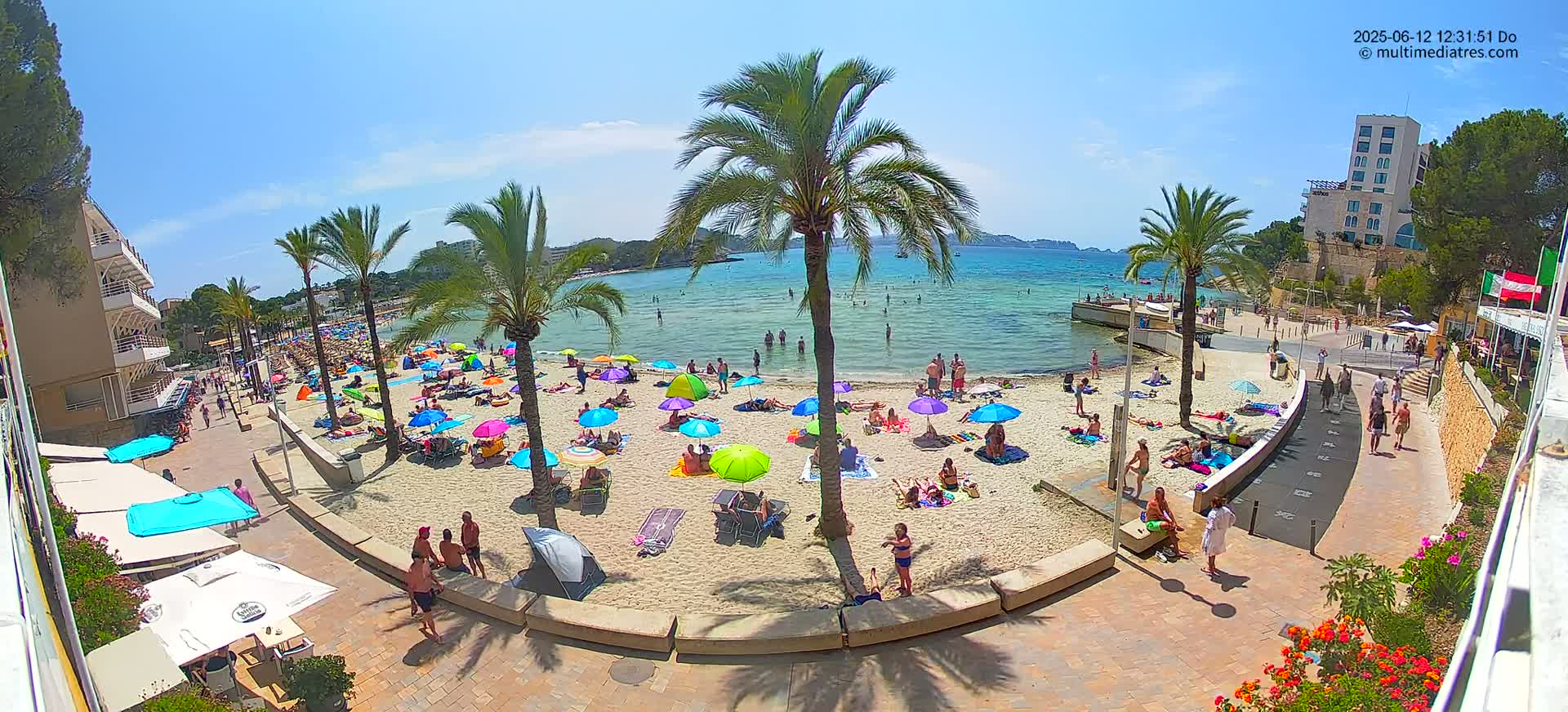 A sunny beach scene shows many people relaxing under colorful umbrellas on a sandy beach next to a calm, clear body of water, with palm trees and buildings in the background.