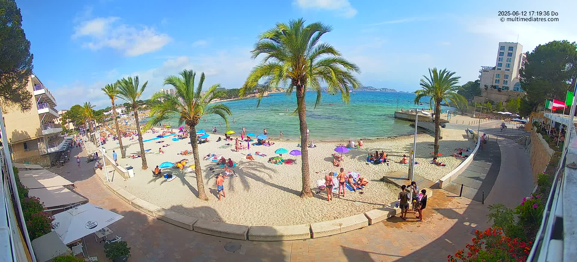 A sunny beach scene shows many people relaxing under umbrellas on a sandy beach, with palm trees and calm turquoise water visible in the background.