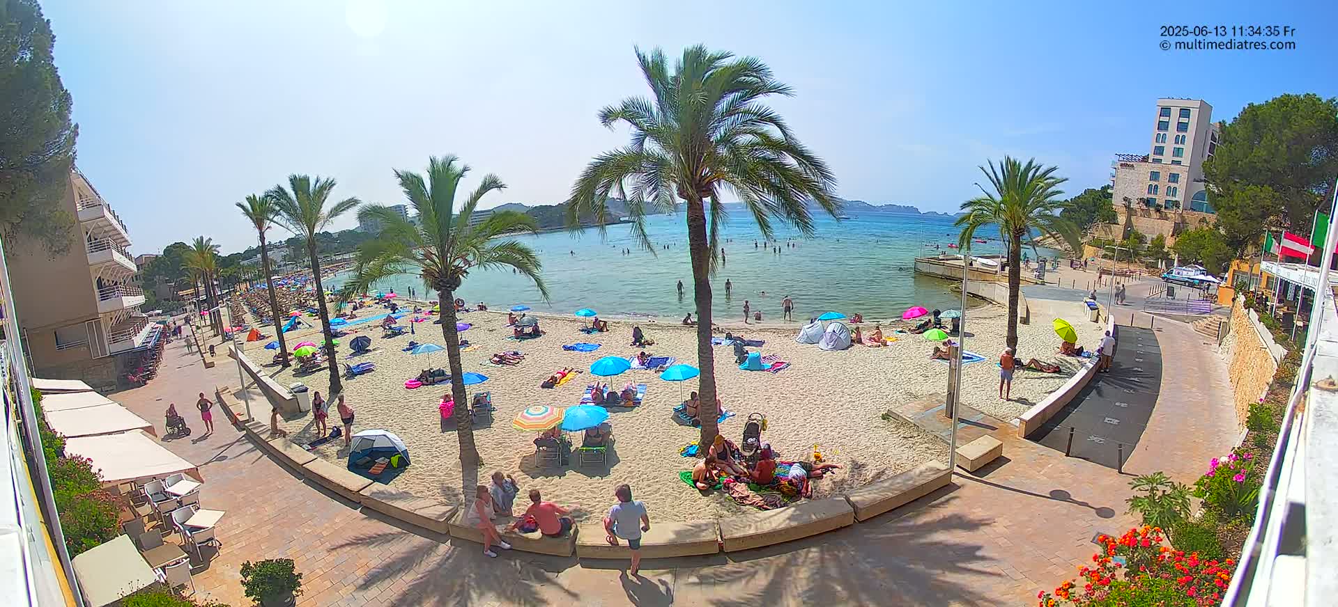 A sunny beach scene shows many people relaxing under umbrellas and in the shallow water, palm trees lining a sandy shore next to a paved walkway and buildings.