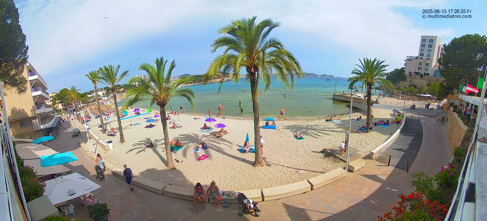 A sandy beach with numerous people sunbathing under umbrellas, palm trees, and buildings is visible on a sunny day.