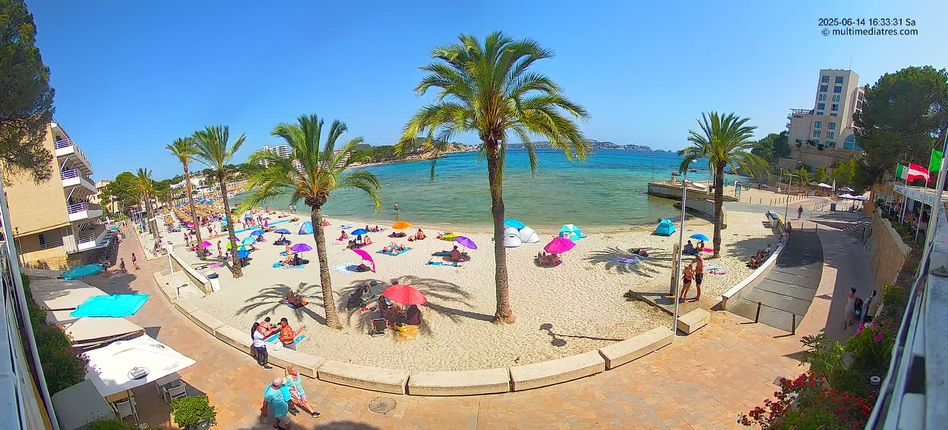 A sunny beach scene shows many people relaxing under colorful umbrellas on a sandy beach next to a calm body of water, palm trees, and buildings.