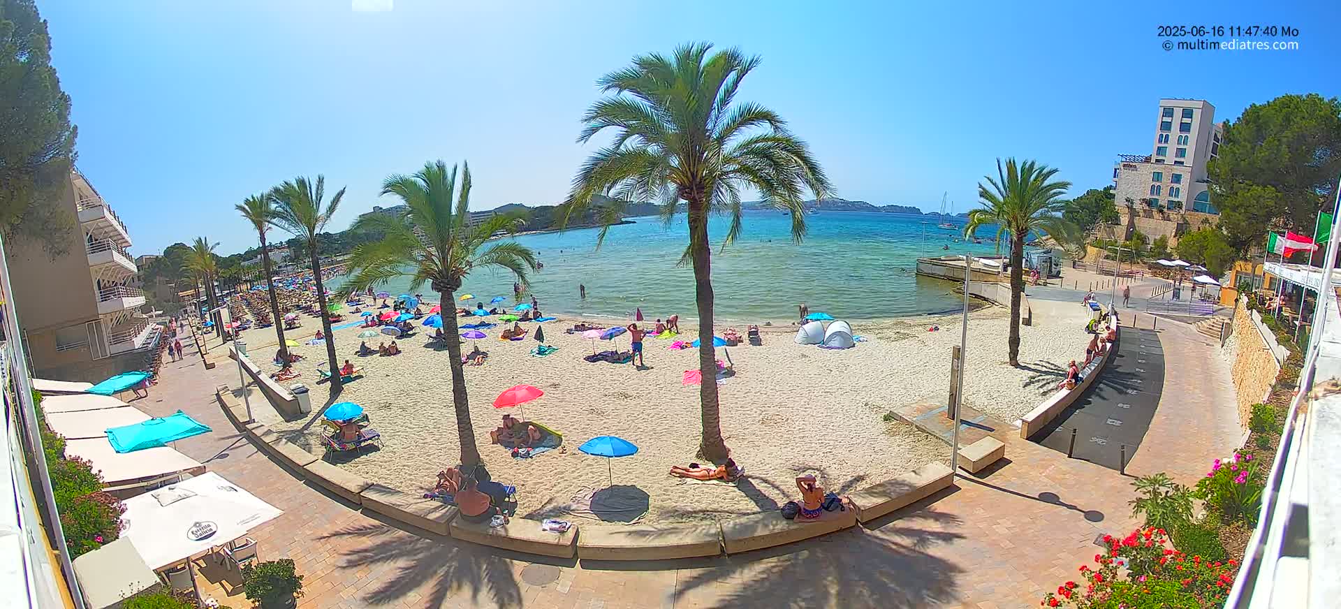 A sunny beach scene shows many people relaxing under umbrellas on a sandy beach next to calm, clear water, with palm trees and buildings visible in the background.