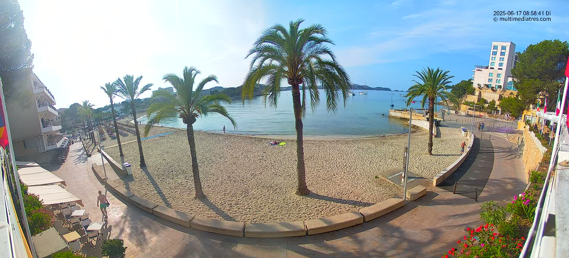 A sandy beach with palm trees and a calm bay under sunny skies is viewed from a high vantage point, showing a paved walkway and buildings nearby.