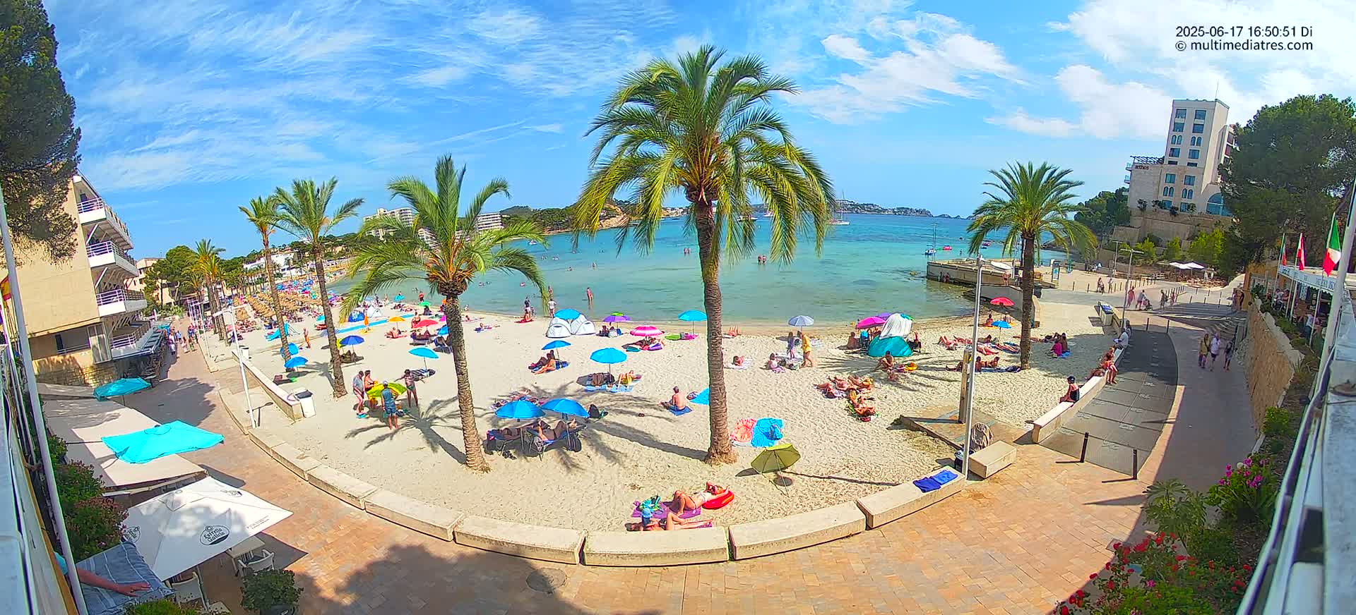 A sunny beach scene shows many people relaxing under colorful umbrellas on a sandy beach next to calm, clear water, with palm trees and buildings in the background.