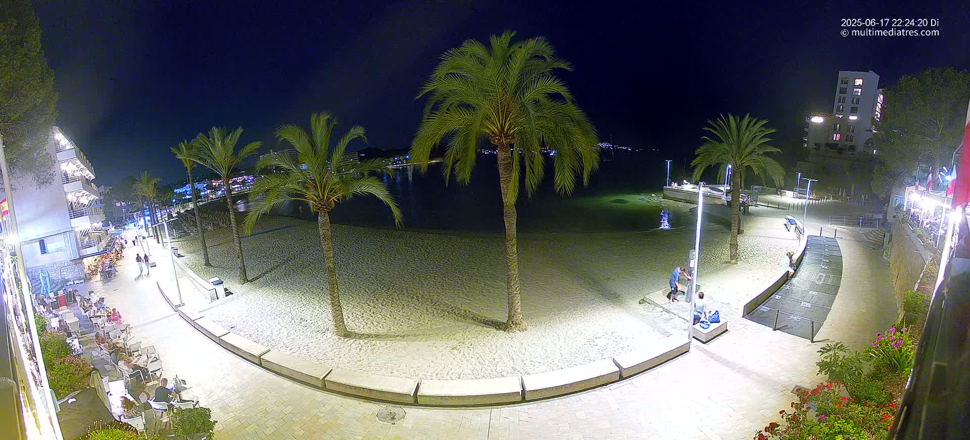 A nighttime view of a sandy beach with palm trees, a calm body of water, and a paved walkway lined with buildings and outdoor seating.