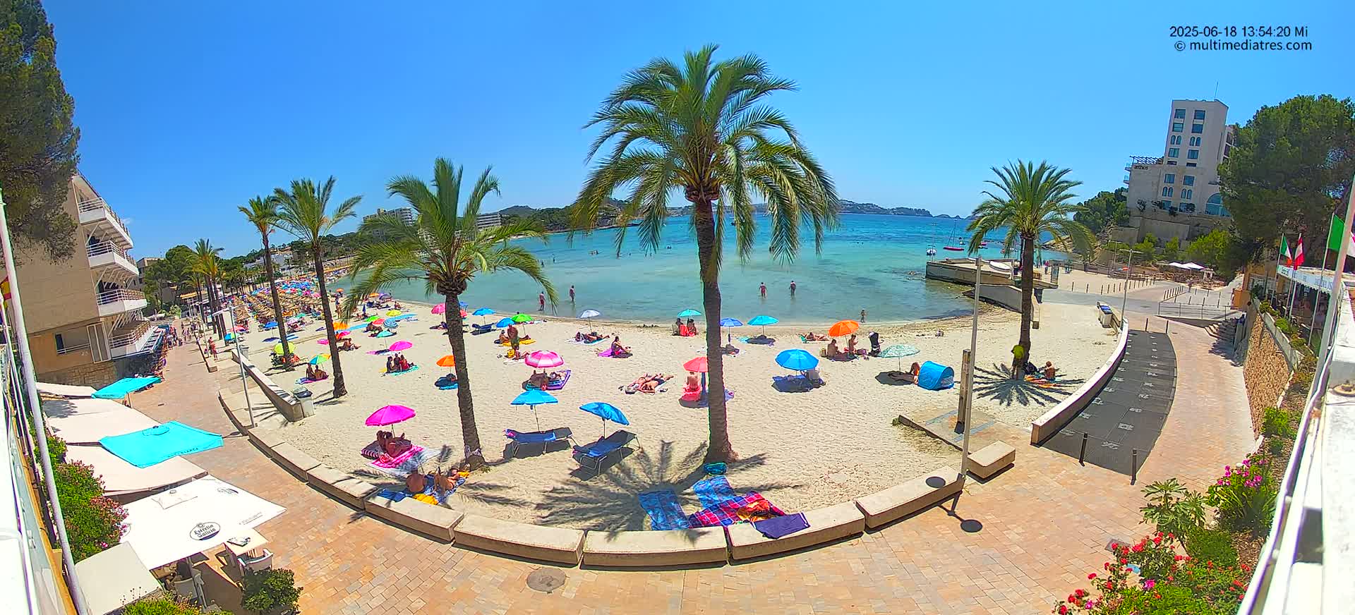 A sunny beach scene shows many people relaxing under colorful umbrellas on a sandy beach next to calm, clear water, with palm trees and buildings in the background.