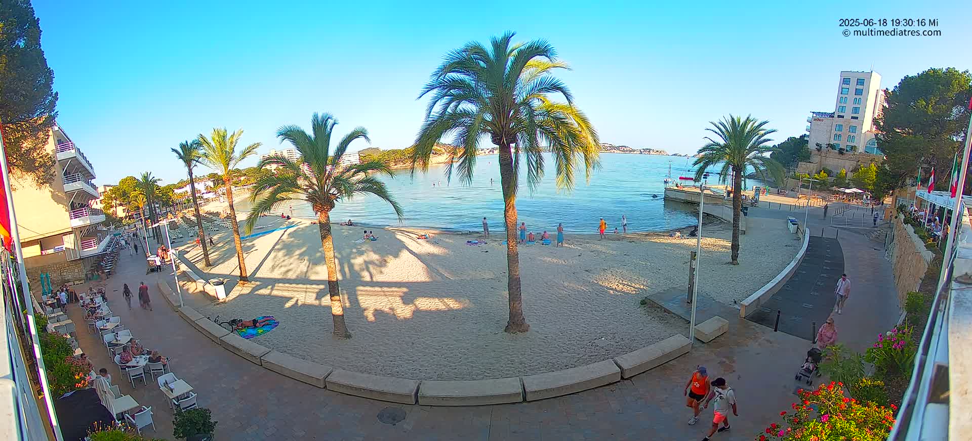 A sandy beach with palm trees and several people is situated next to a calm, clear body of water under a sunny sky, with buildings and a paved walkway visible nearby.