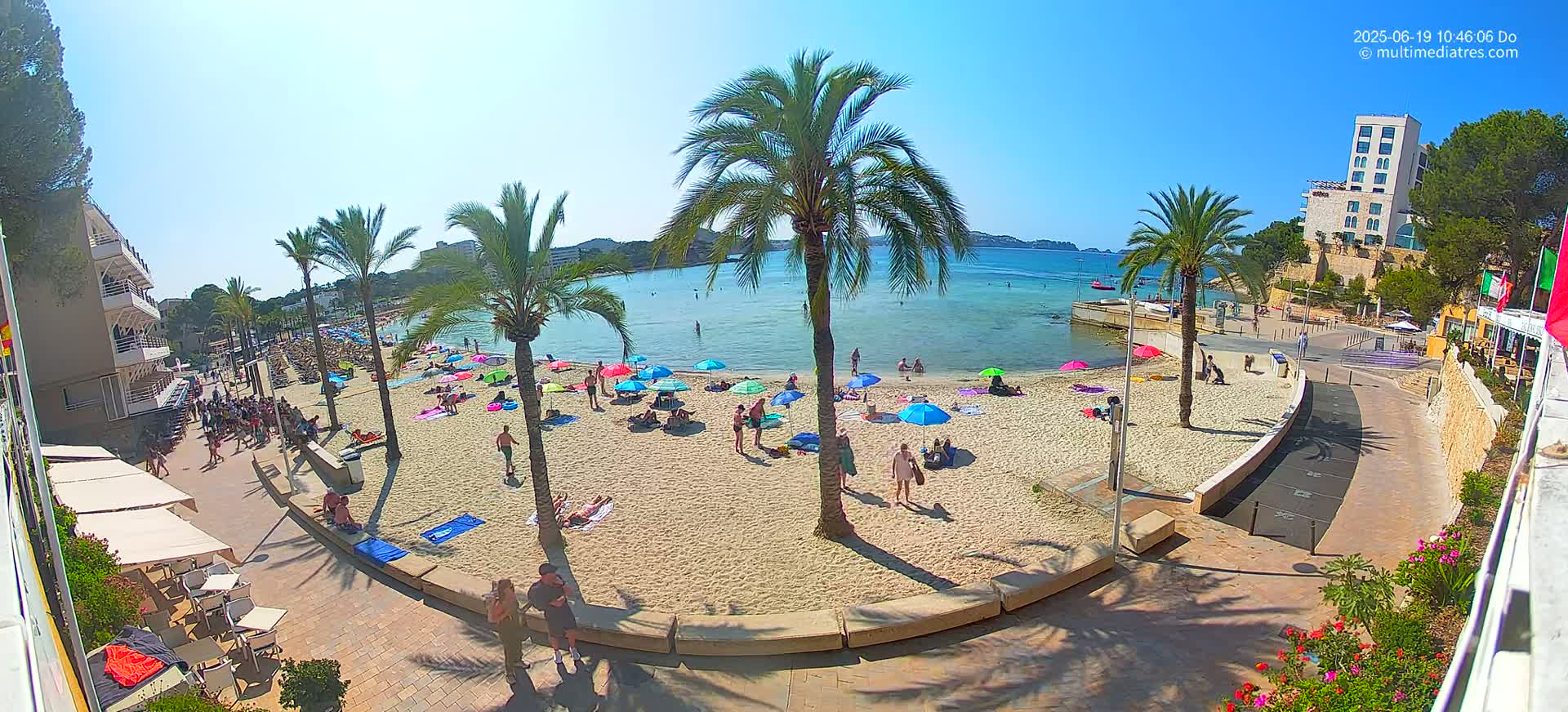 A sunny beach scene shows people relaxing under colorful umbrellas on a sandy beach next to calm, clear water, with palm trees and buildings in the background.