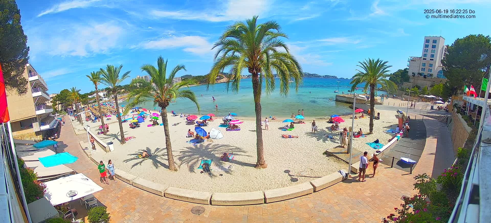 A sandy beach with people relaxing under colorful umbrellas, palm trees, and buildings, on a sunny day with clear blue skies.