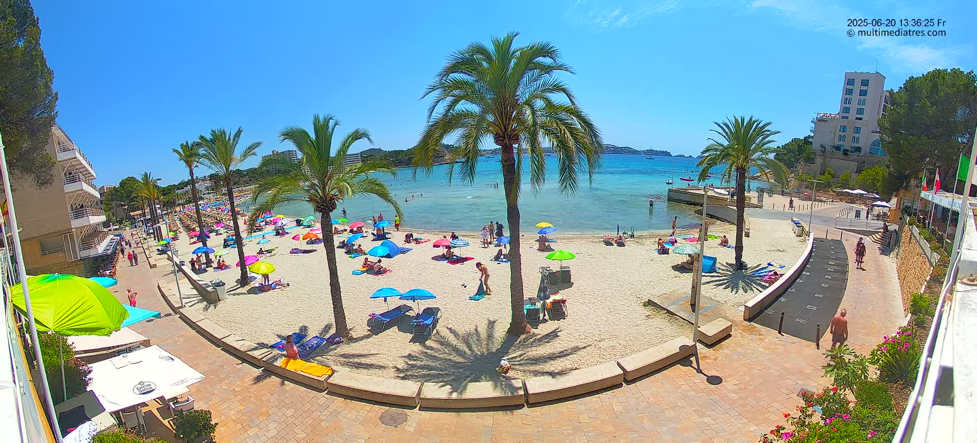 A sandy beach with colorful umbrellas and sunbathers is bordered by palm trees, buildings, and a paved walkway under a bright sunny sky.