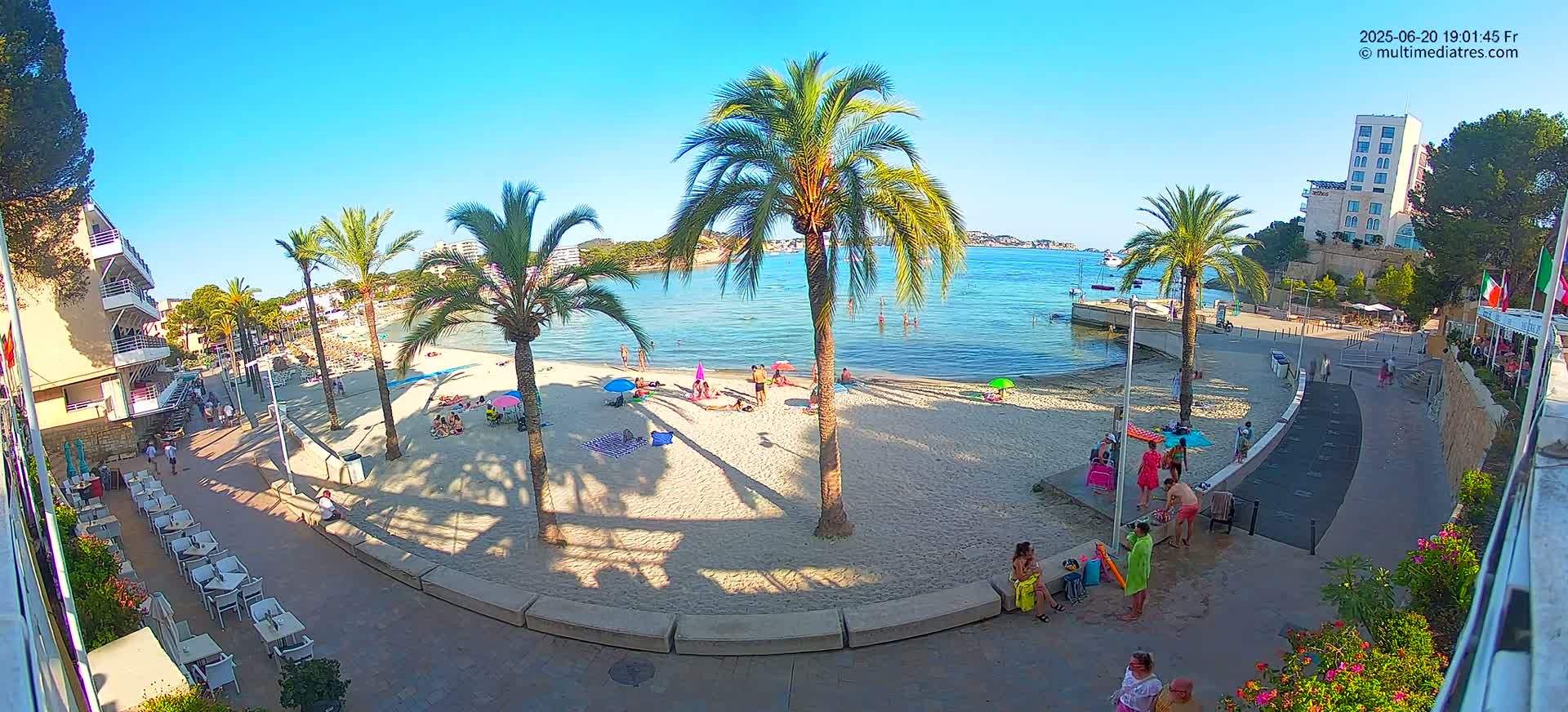 A sunny beach scene shows people relaxing on the sand and swimming in the calm, clear water, with palm trees and buildings lining the shore.