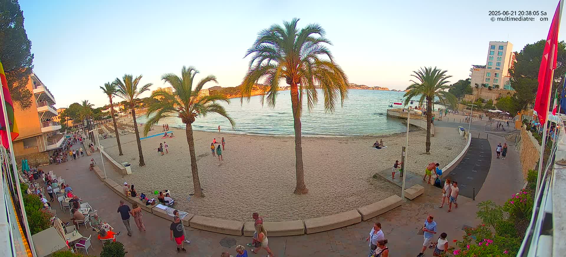 A sandy beach with palm trees, people relaxing and walking along a paved walkway, overlooking calm water at sunset.