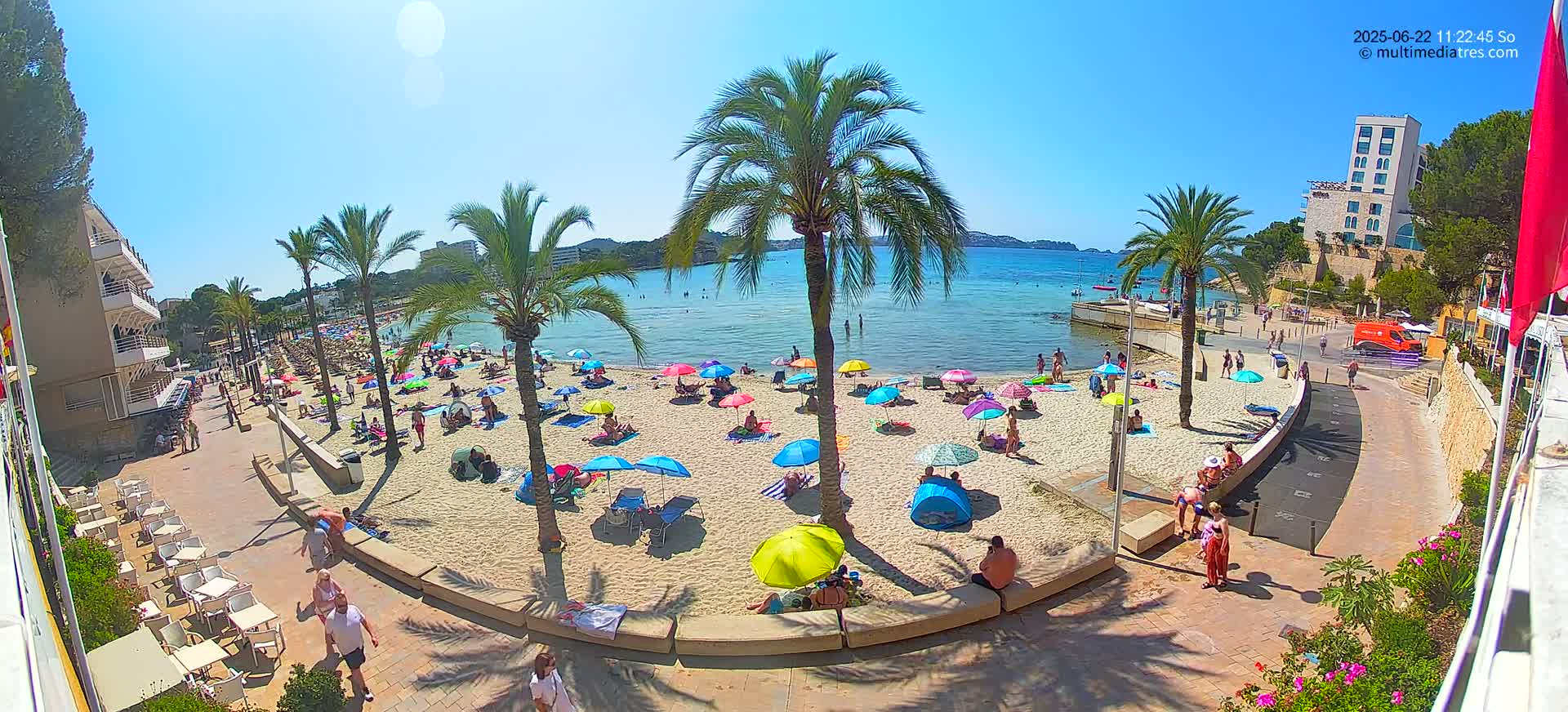 A sunny beach scene with numerous colorful umbrellas and people relaxing on the sand, palm trees lining the shore, and a calm body of water in the background.