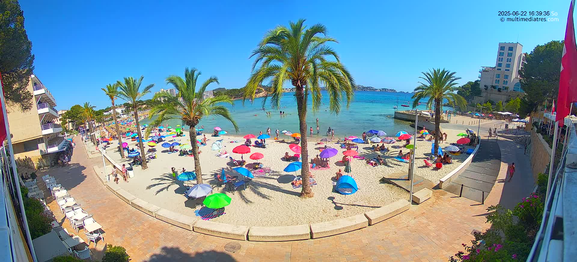 A sunny beach scene shows numerous colorful umbrellas and people relaxing on a sandy beach next to calm, clear water, with palm trees and buildings lining the shore.