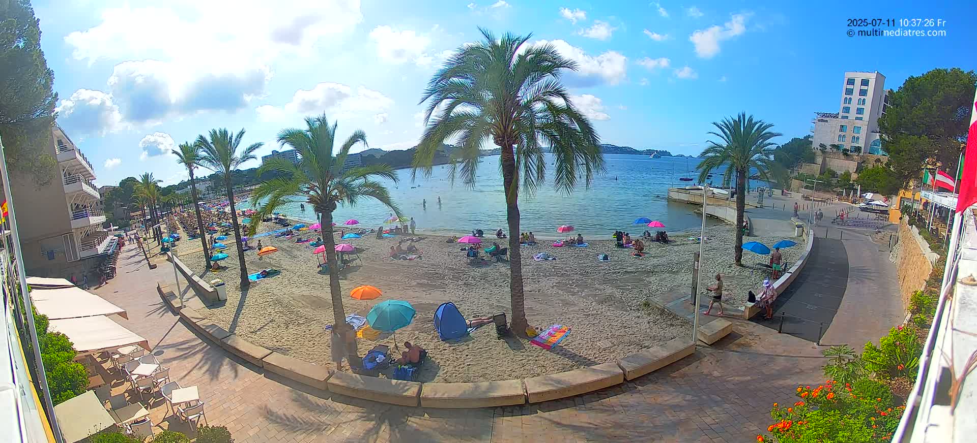 A sandy beach with palm trees and colorful umbrellas, under a partly sunny sky, is bordered by a paved walkway and buildings.