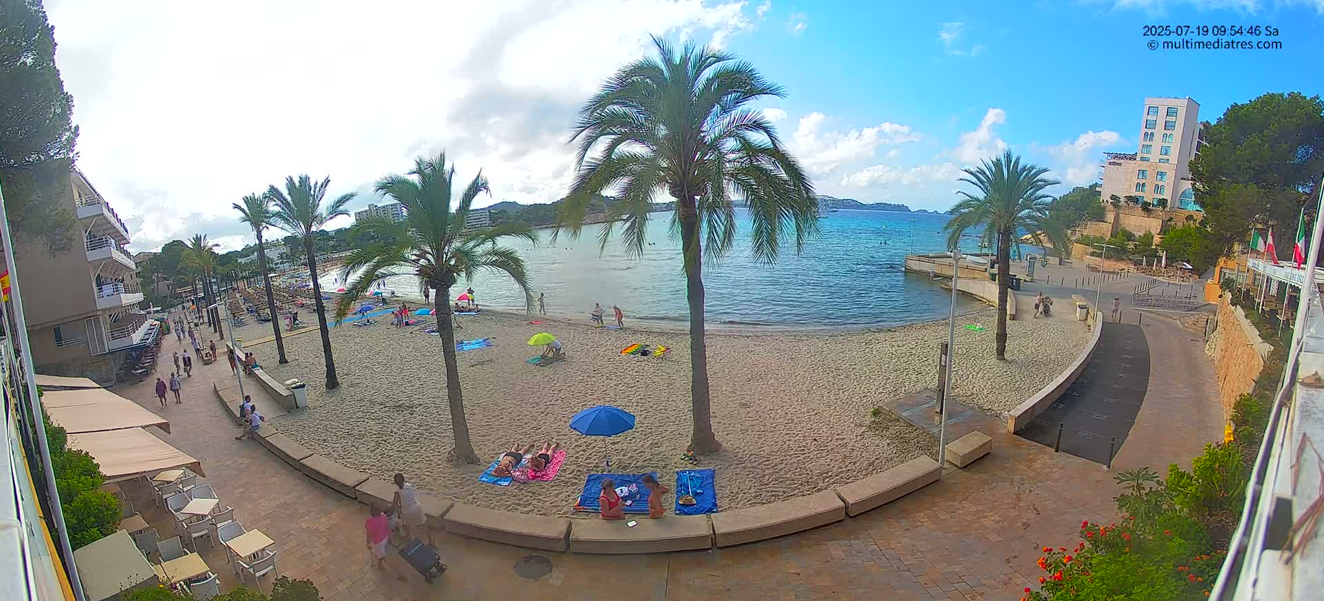 A sandy beach with palm trees and several people relaxing under umbrellas, is adjacent to a paved walkway and buildings, under a partly cloudy sky.