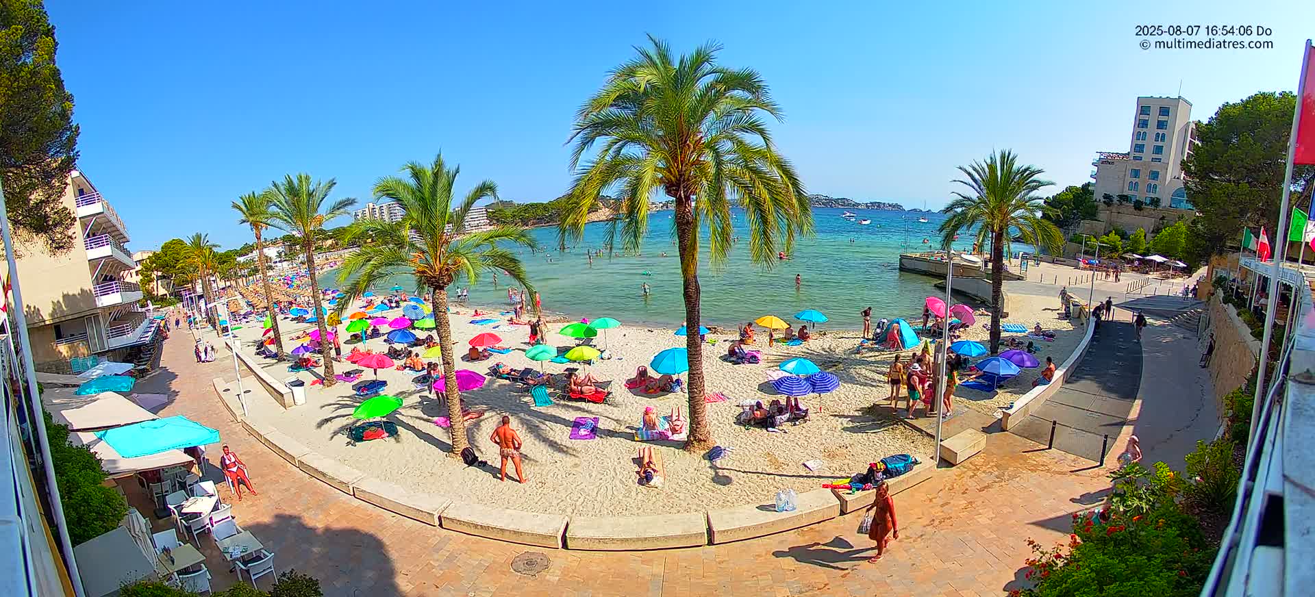 A sunny beach scene shows many people relaxing under colorful umbrellas on a sandy beach next to calm, clear water, with palm trees and buildings in the background.