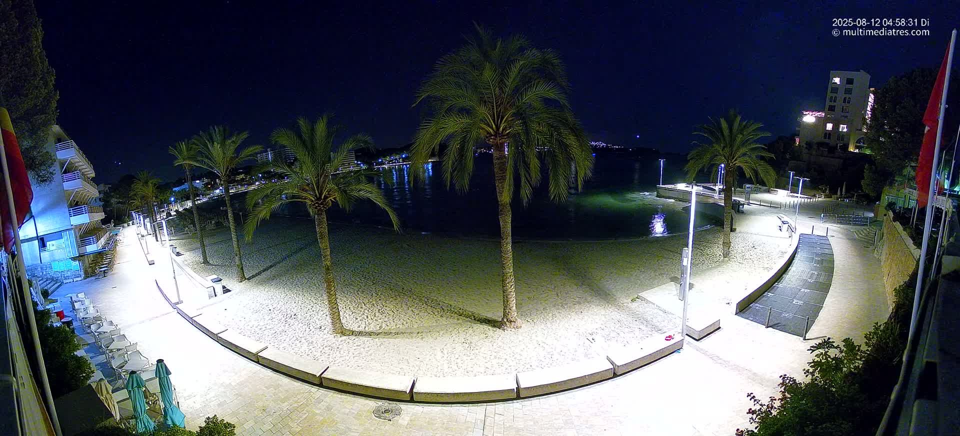 A nighttime, high-angle view shows a sandy beach bordered by a paved walkway lined with palm trees and lit by lampposts, with calm water visible beyond.