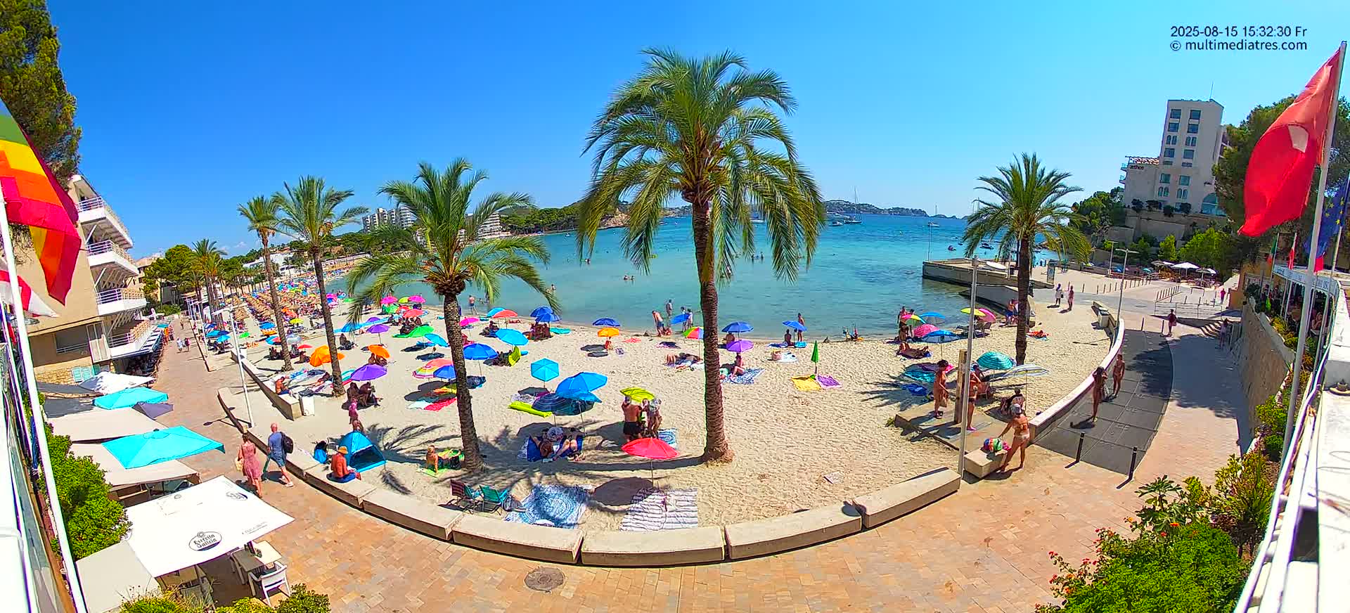 A sandy beach with palm trees, beach umbrellas, and people enjoying a sunny day by calm blue water, next to buildings and a paved walkway.