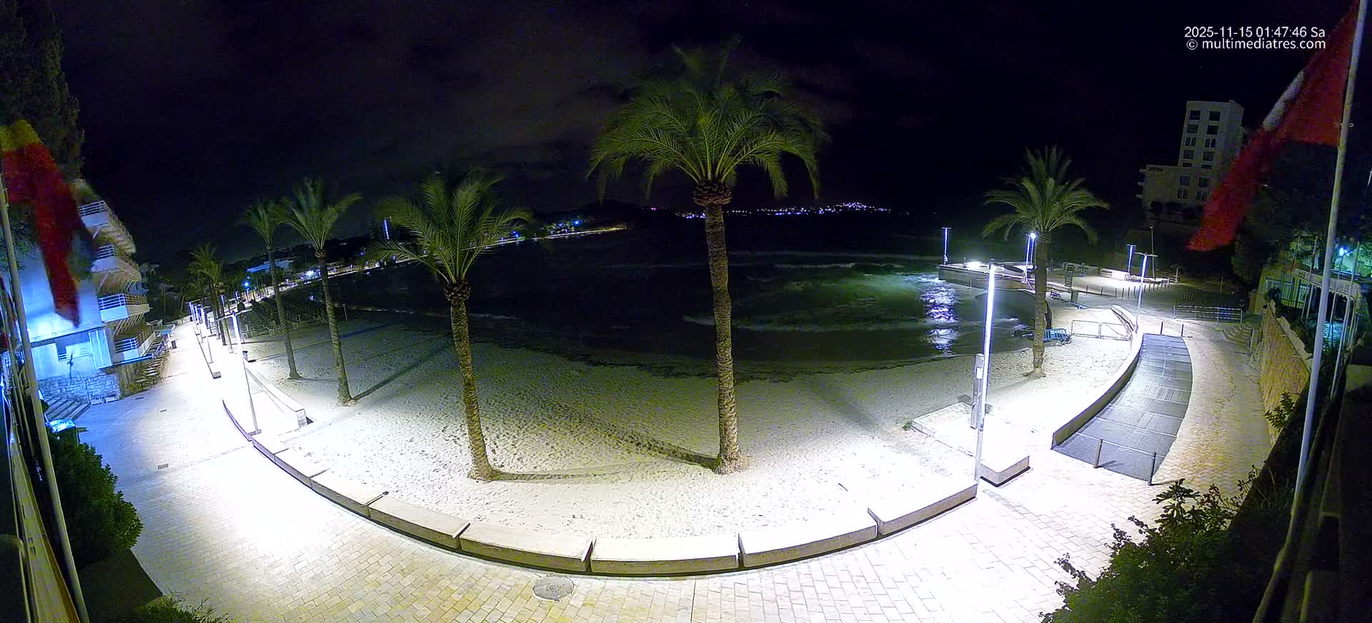 A wide-angle nighttime view shows a brightly lit sandy beach lined with palm trees and modern streetlights, leading to a calm bay with distant city lights under a dark, cloudy sky.
