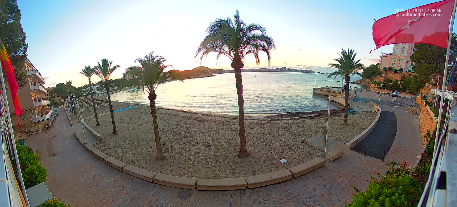 A wide-angle nighttime view shows a brightly lit sandy beach lined with palm trees and modern streetlights, leading to a calm bay with distant city lights under a dark, cloudy sky.