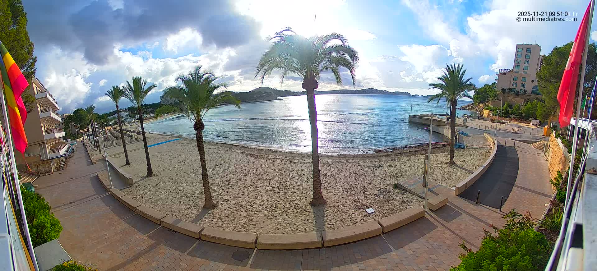 On a sunny, partly cloudy morning, a tranquil sandy beach with several palm trees and a paved promenade stretches along a sparkling blue bay, backed by various coastal buildings.